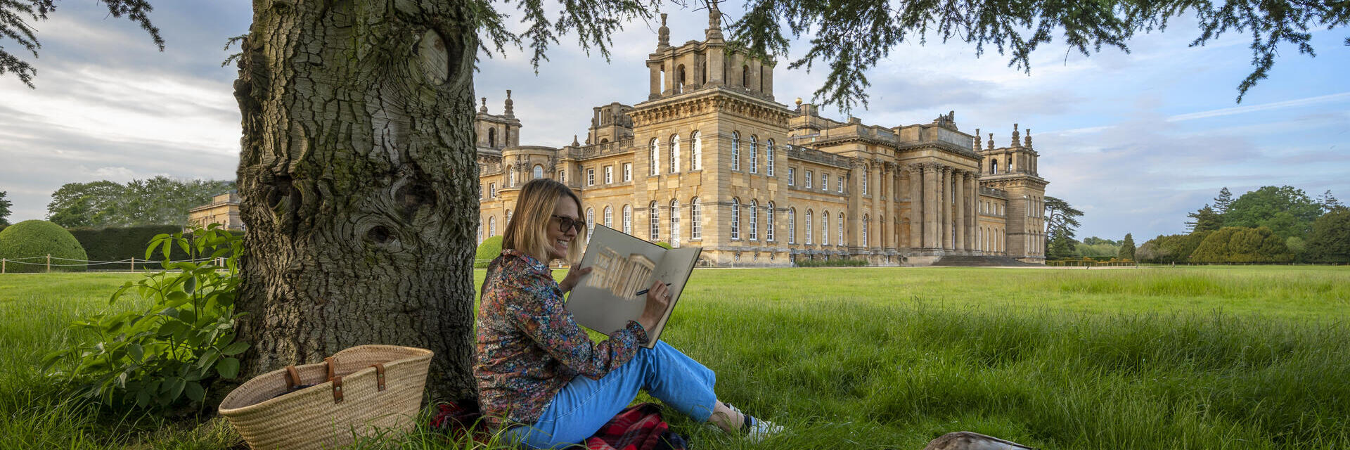 Frau sitzt unter einem Baum und zeichnet auf dem Gelände eines großen Landguts