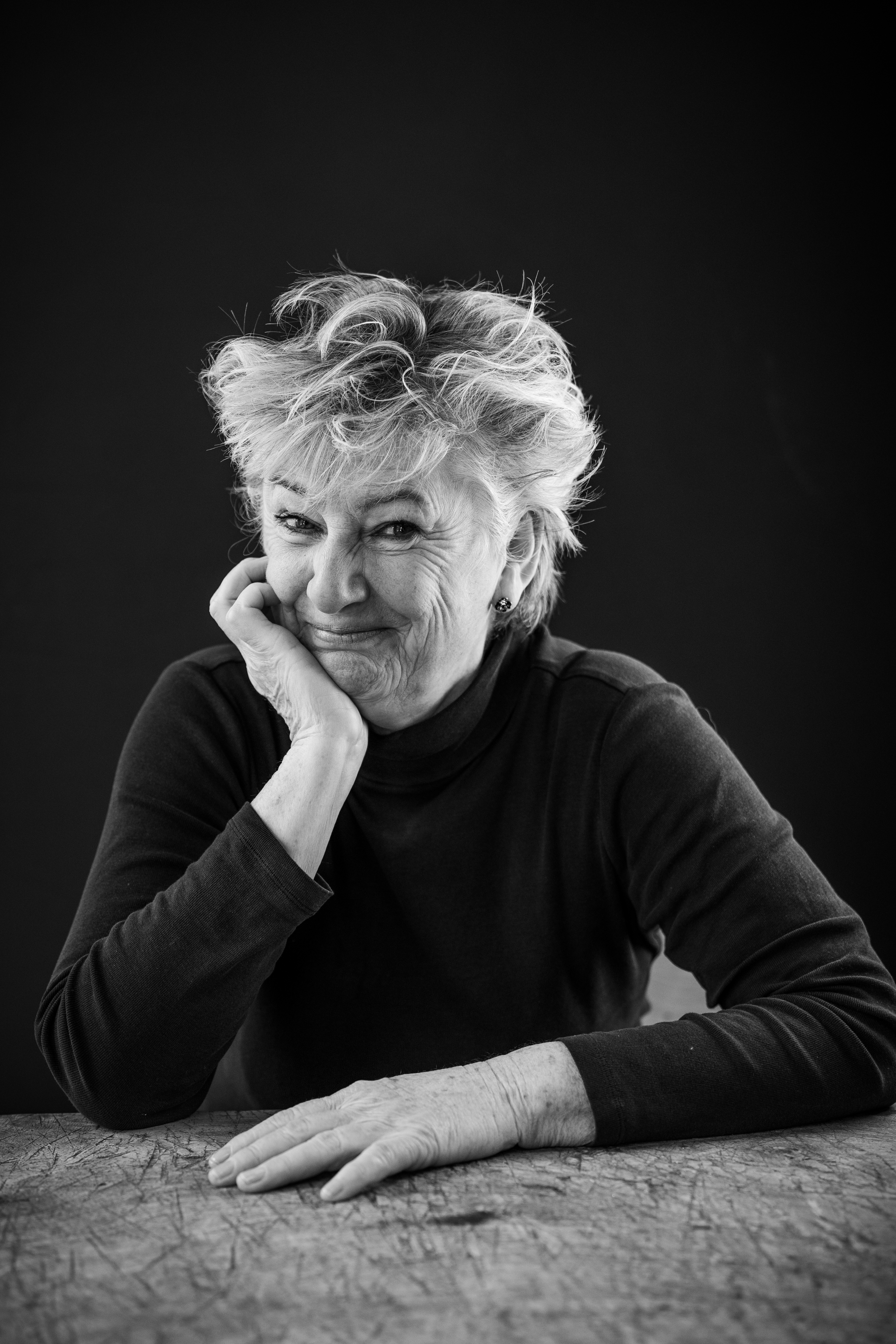 Black and white portrait of an older person smiling, seated at a table, with a dark background.