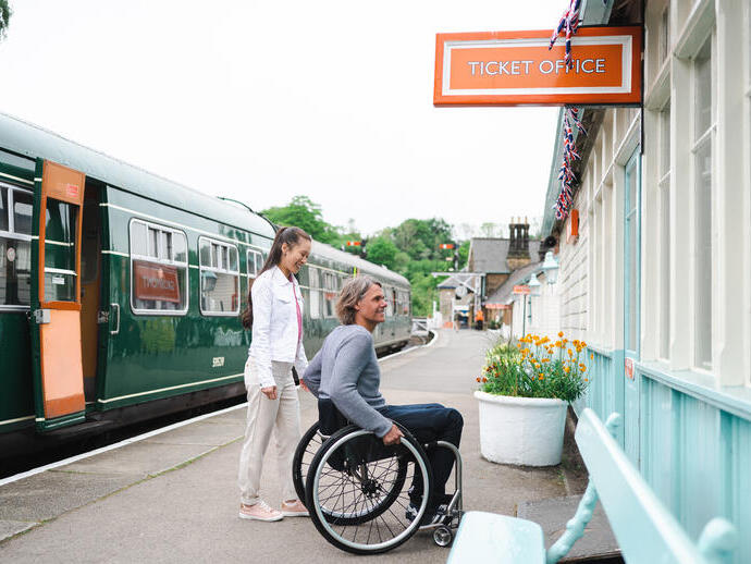 Man using a wheelchair and woman about to go into the ticket office at a station.