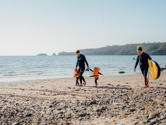 عائلة تمشي على الشاطئ، الرجل بصحبة طفلين صغيرين والمرأة تحمل لوح تزلج على شاطئ سوندرسفوت