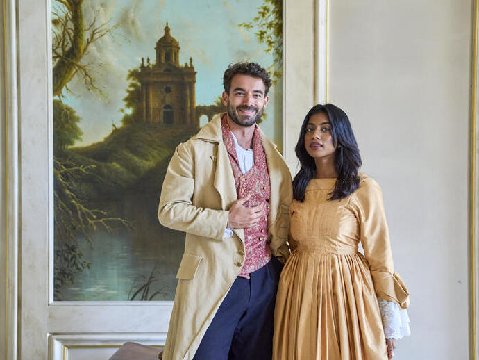 Ein Mann und eine Frau in historischen Kostümen stehen in einem dekorierten Raum vor einem Gemälde mit Landschaft und altem Gebäude.