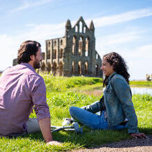 Man and woman sitting on grass with crutches beside them, an Abbey in the background