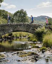 Un grupo de personas camina sobre un puente de piedra sobre un río, rodeado de paisaje verde y colinas bajo un cielo soleado.