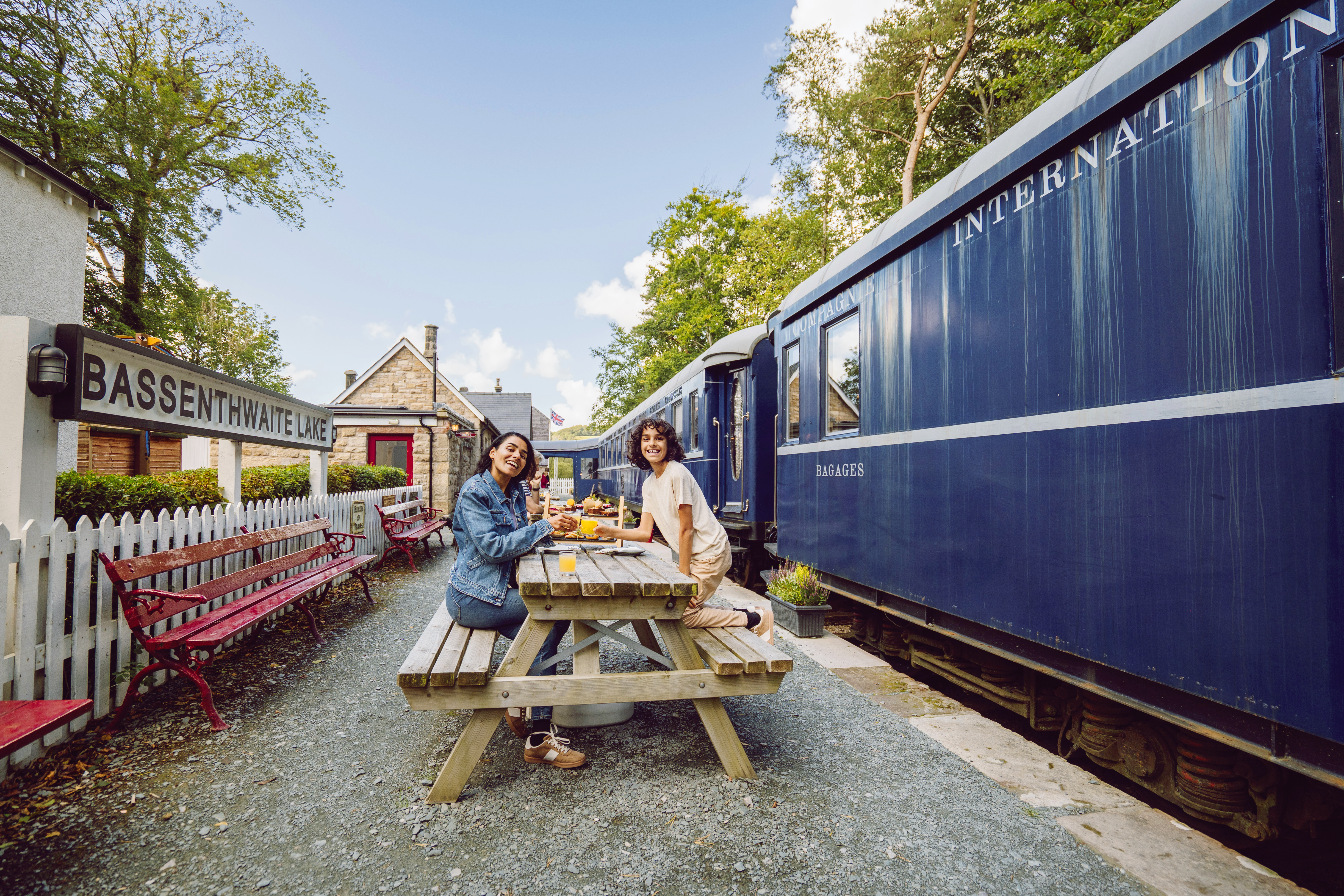 Two people dining at a picnic table on a historic train station platform with a blue train and Bassenthwaite Lake station sign visible.