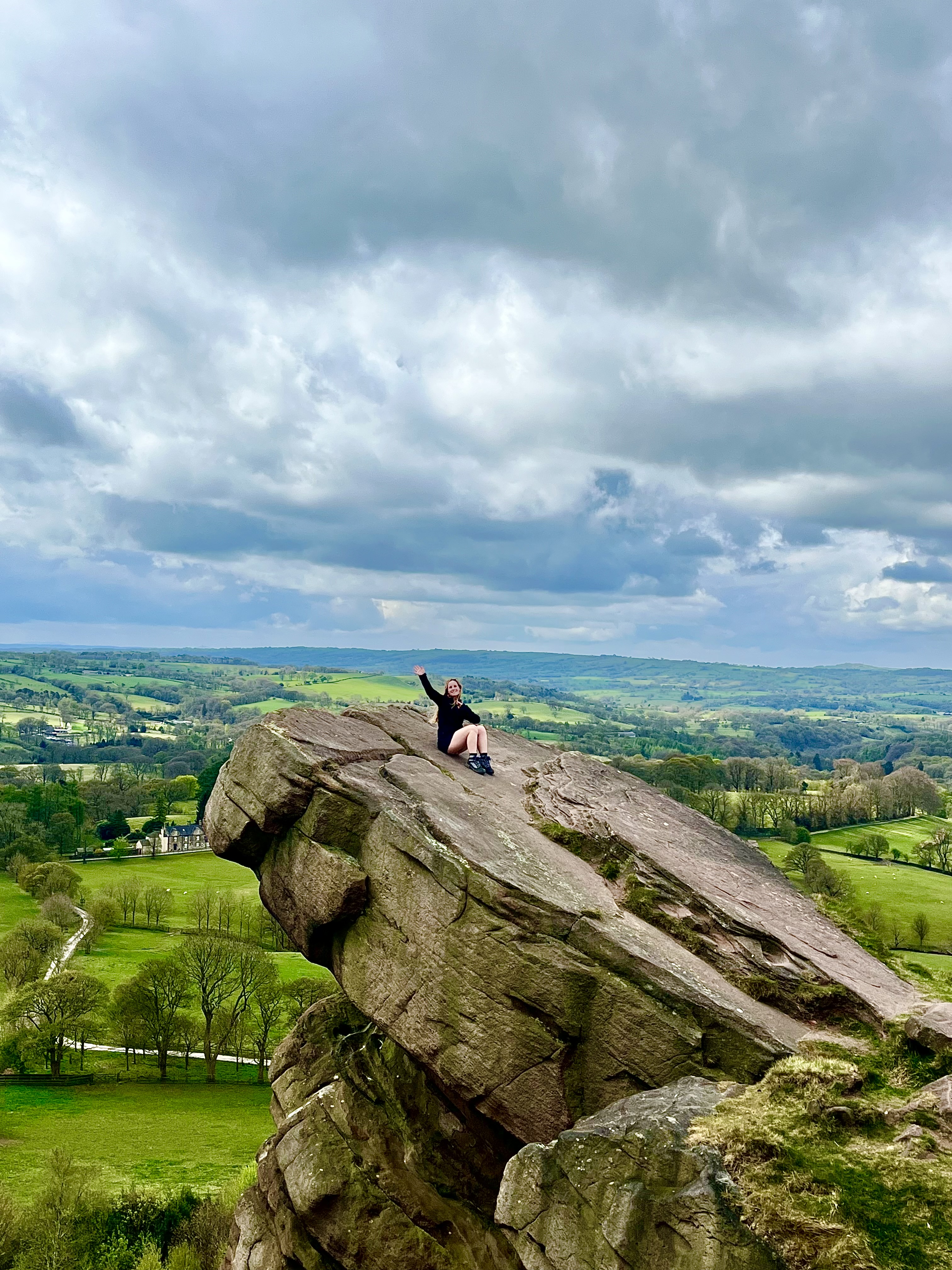 Person sits on large rocky outcrop with scenic countryside and dramatic cloudy sky in the background.