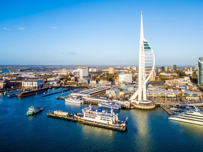 A waterfront cityscape featuring a tall, modern tower, ferries, yachts, and urban buildings under a clear blue sky.