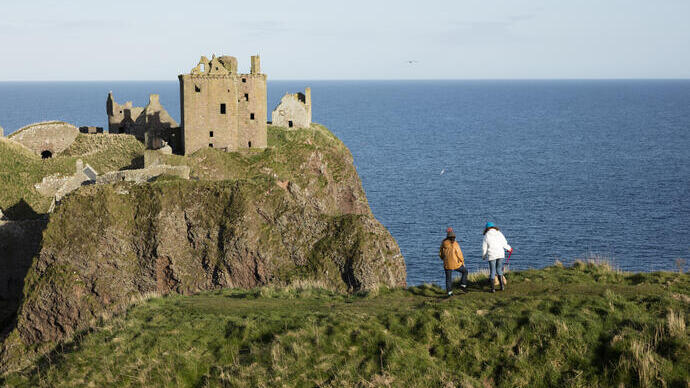 Two women stood on a clifftop, near a castle and the sea