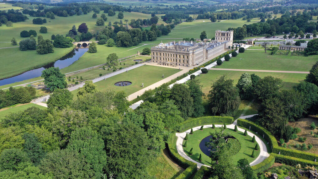 Aerial view of a grand historic estate with formal gardens, a river, a stone bridge, and rolling green landscape.
