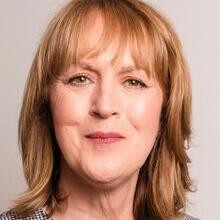 Portrait of a middle-aged woman with shoulder-length light brown hair, wearing a black and white patterned top, smiling slightly, with a plain background.