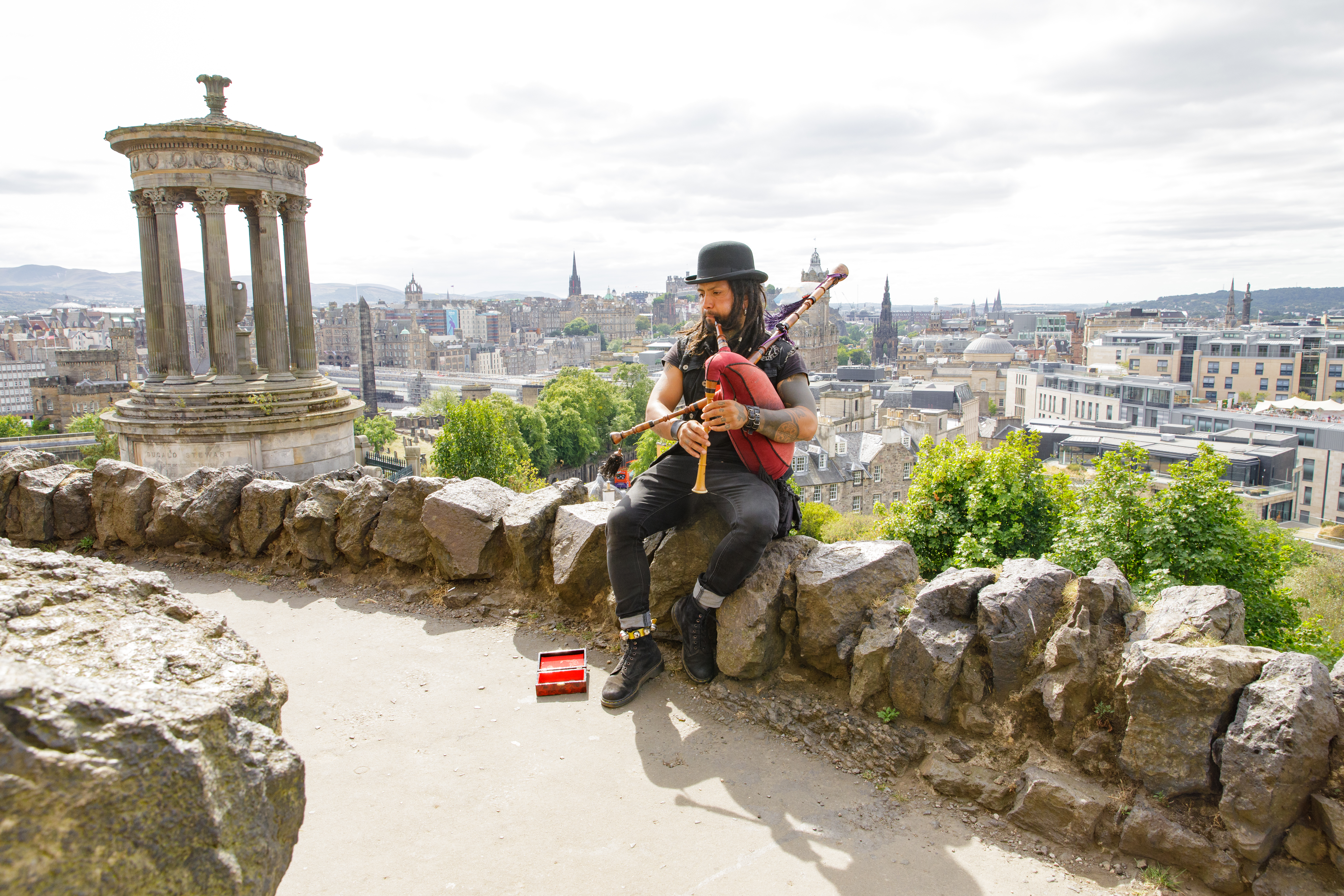 Dudelsackspieler sitzt auf einer Steinmauer mit Stadtblick und klassischem historischen Denkmal links.