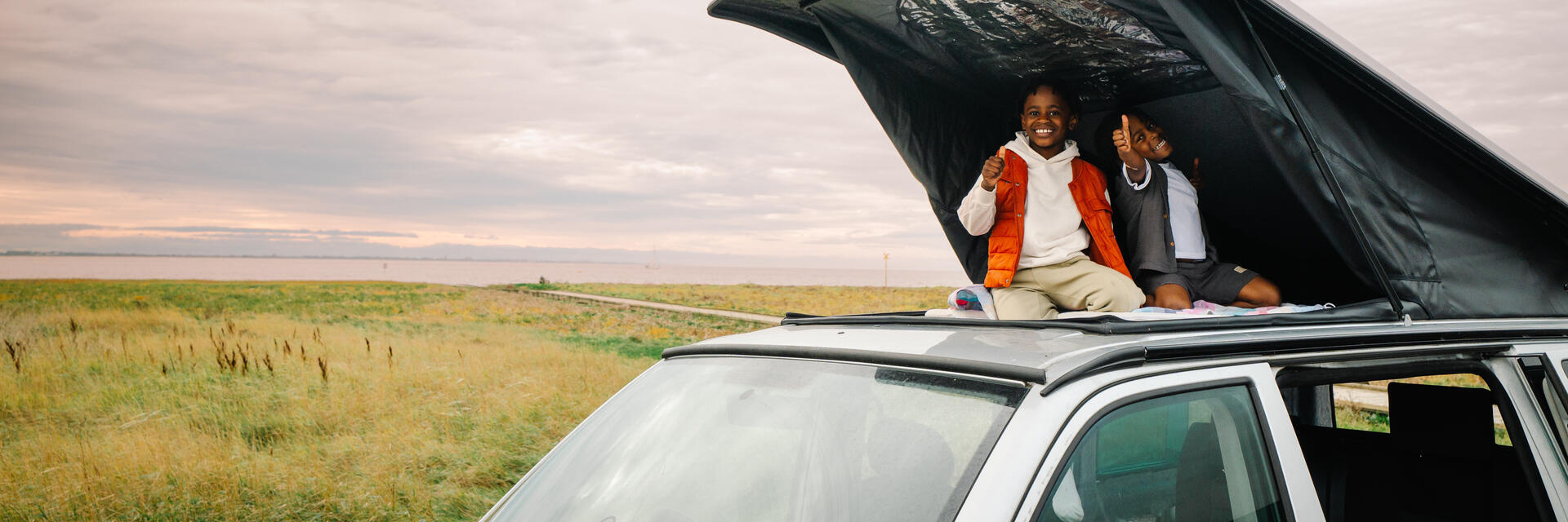 Two children sitting and smiling in a rooftop tent on a car, with grassland and water in the background under a cloudy sky.