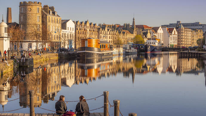 Two young women sat on the jetty of the waterfront in the evening with boats and buildings by the harbour.