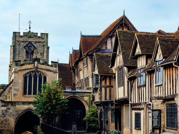 Narrow path beside old timber buildings leading to black gates and a church in the distance