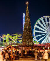 A Christmas fair at night with a large decorated Christmas tree, ferris wheel, roller coaster, lights, and crowds of people.