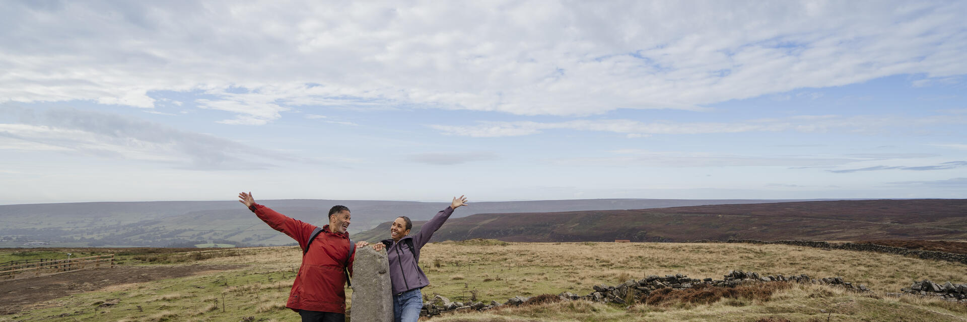 Two people posing cheerfully beside a standing stone in open countryside under a wide, partly cloudy sky. Rolling hills and grassy moorland in the background.