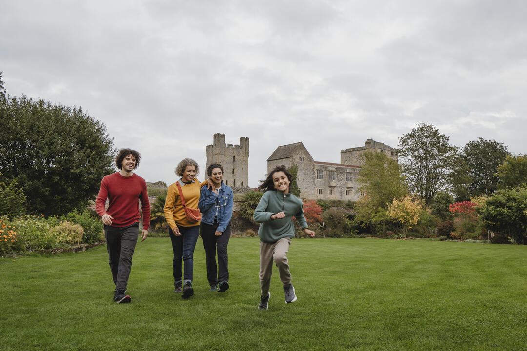 Four people walking on a large green lawn with an old stone castle and autumn trees in the background, under a cloudy sky.