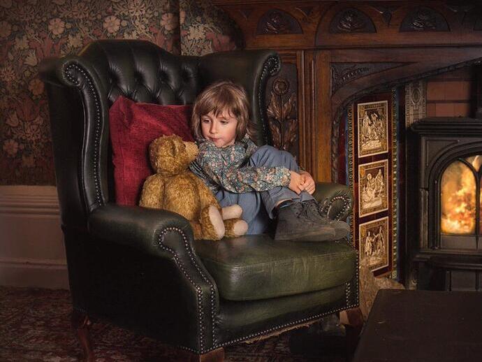 Young child sat on a green armchair in front of a fireplace with a teddy bear next to them at Criag y Nos Castle in the Brecon Beacons