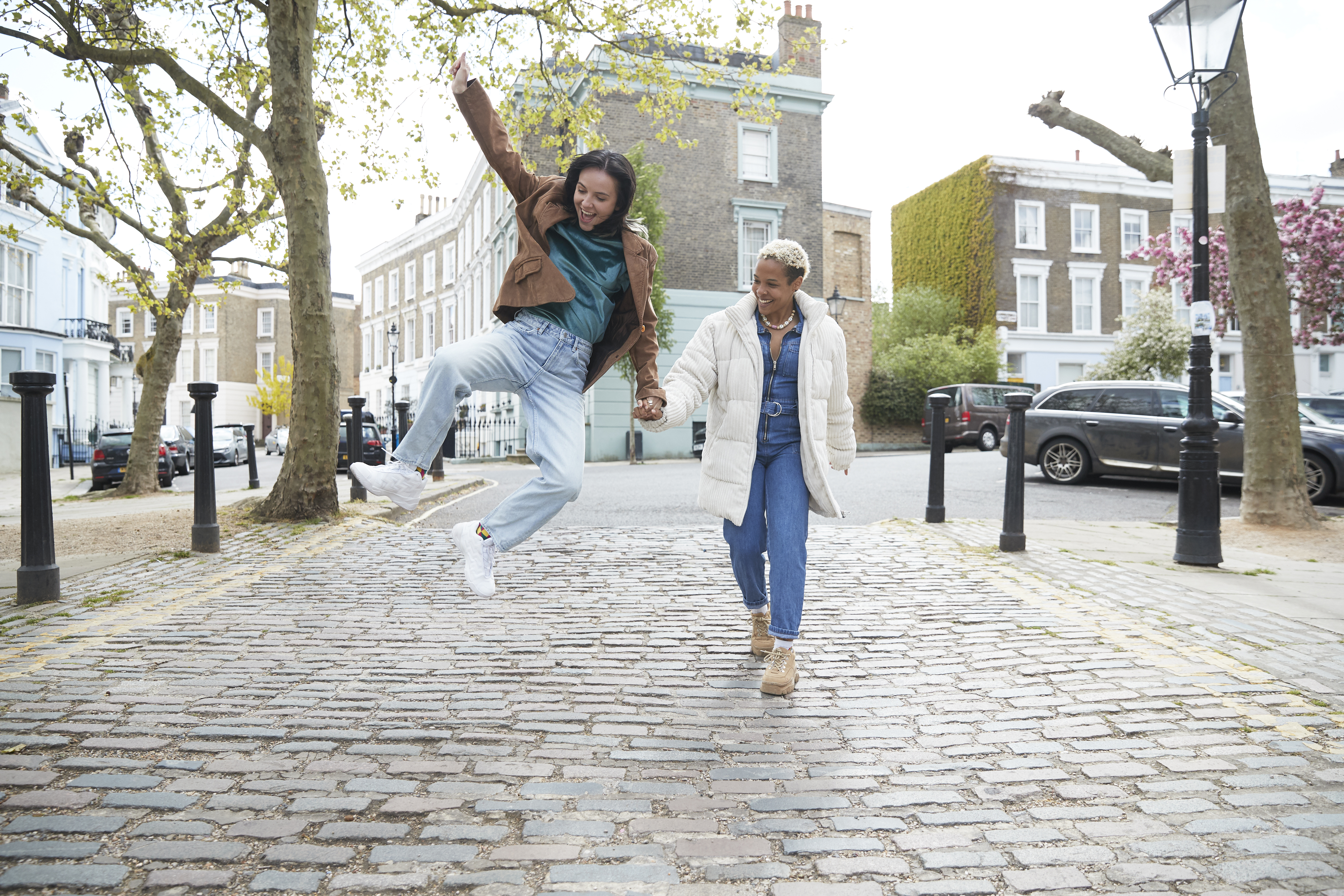 Happy young woman jumping while holding girlfriend's hand walking on a footpath in a street