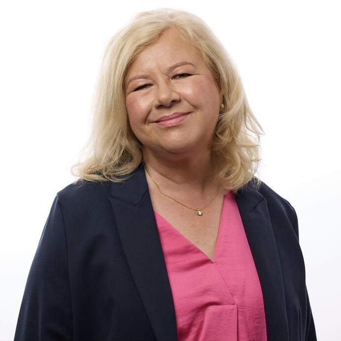 A person with blonde hair smiles at the camera, wearing a dark blazer and pink top, standing against a plain white background.