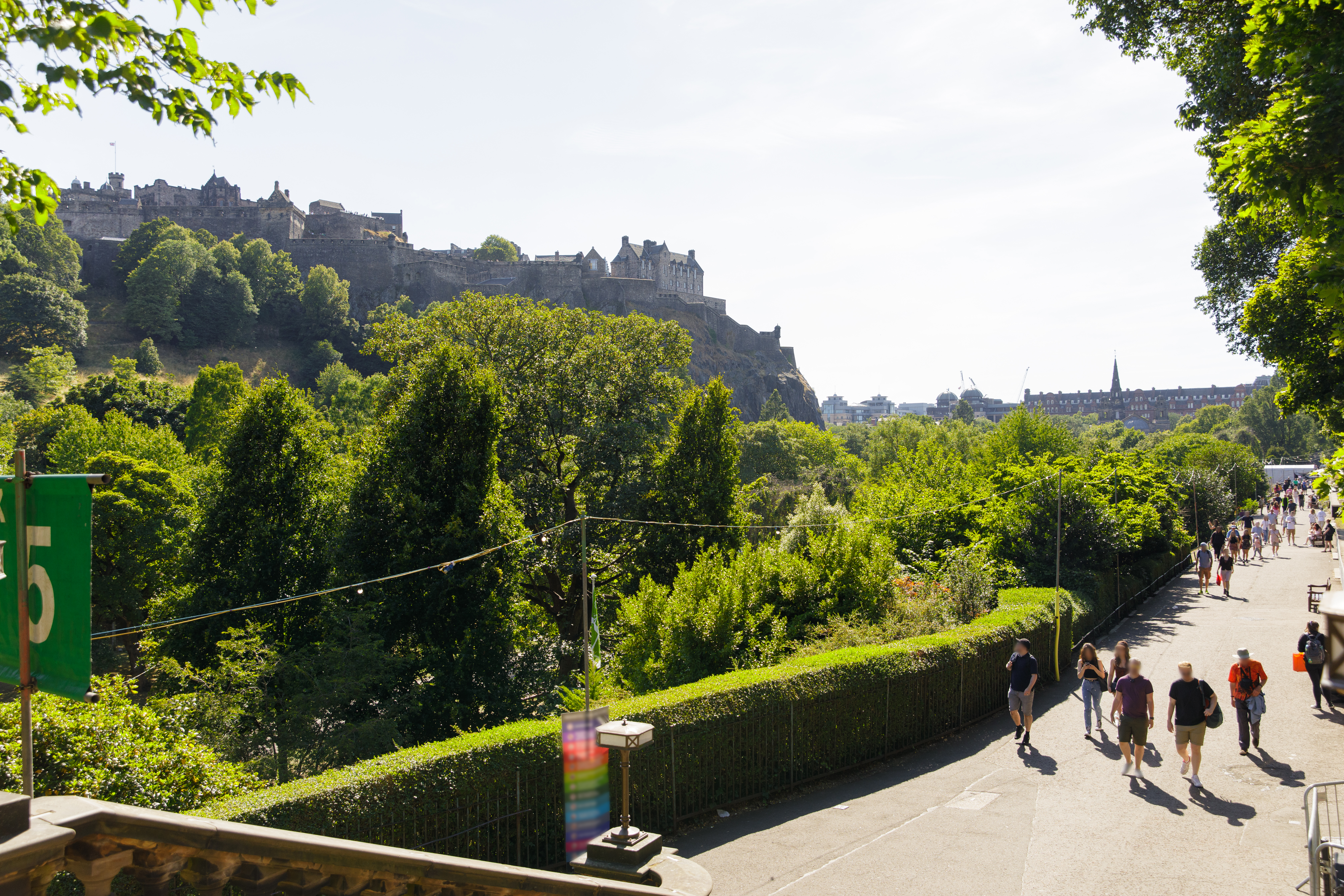 View of Edinburgh Castle surrounded by trees, with people walking along a sunny pathway in the foreground.