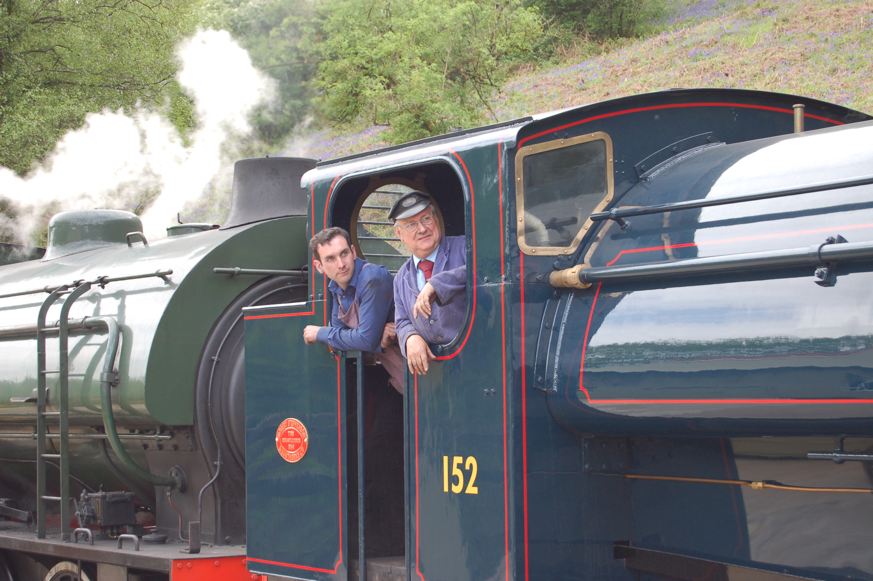 Two men wearing overalls standing in the cab of a vintage steam train.