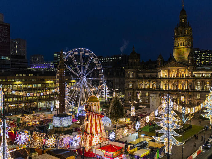 A festive night scene featuring a brightly lit Ferris wheel, holiday market, illuminated decorations, and a historic building in the background.