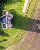 Luftaufnahme eines Backsteingebäudes mit drei Türmen auf einer grünen Wiese, mit langem Schatten aufs angrenzende Feld.