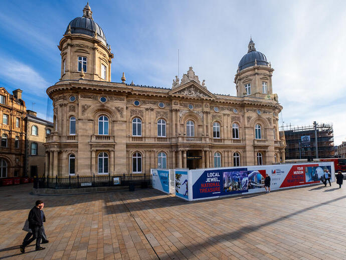 Historic domed building with ornate architecture, plaza in foreground, temporary museum construction signs, and people walking nearby on a sunny day.
