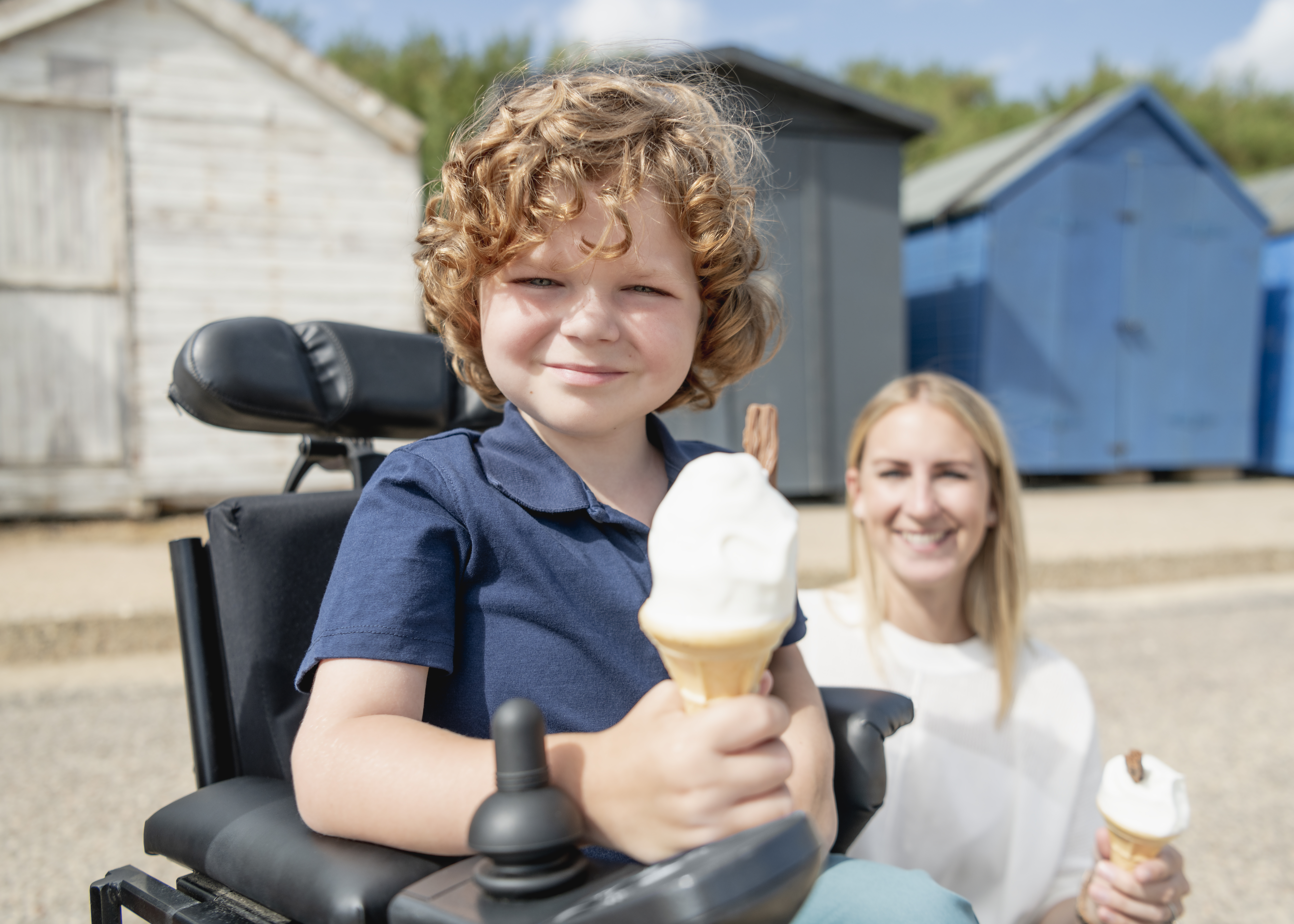 Young electric wheelchair user holding an ice cream cone with mum in background at the seaside
