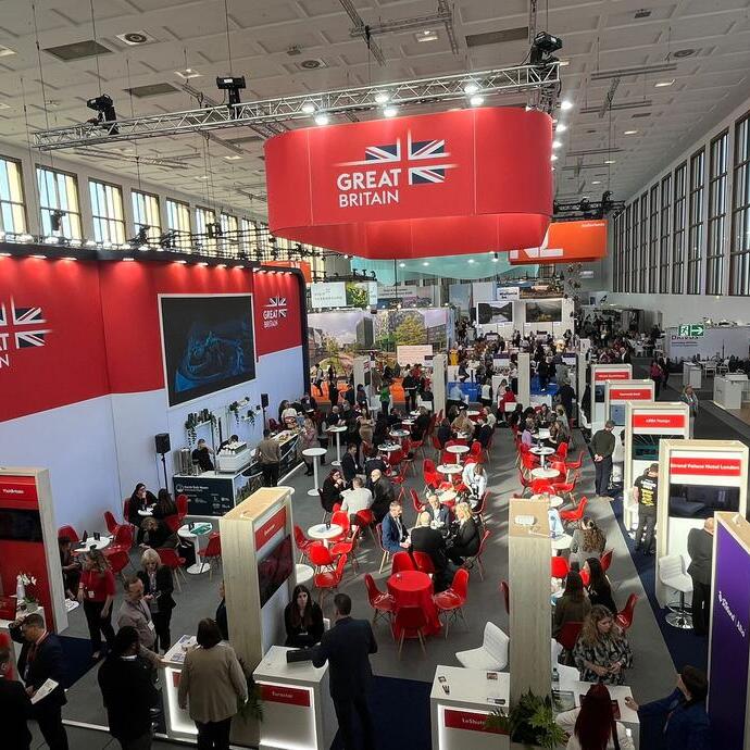 Busy trade show or exhibition hall with red and white Great Britain banners, people networking at tables, and various booths in a modern, bright venue.