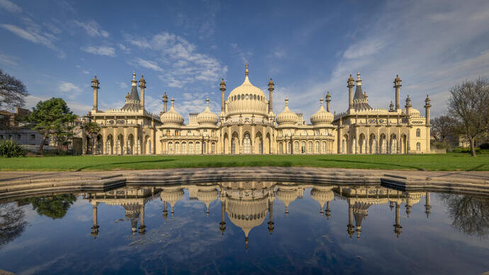 The Royal Pavilion in Brighton with domed roofs and minarets, reflected in a pond under a blue sky and surrounded by green lawns.