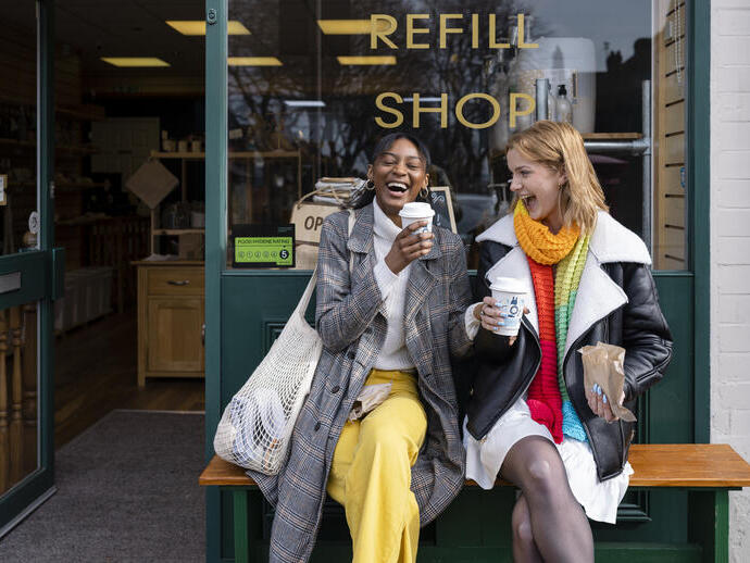 Two young women sat on a bench outside a coffee shop with takeaway coffees