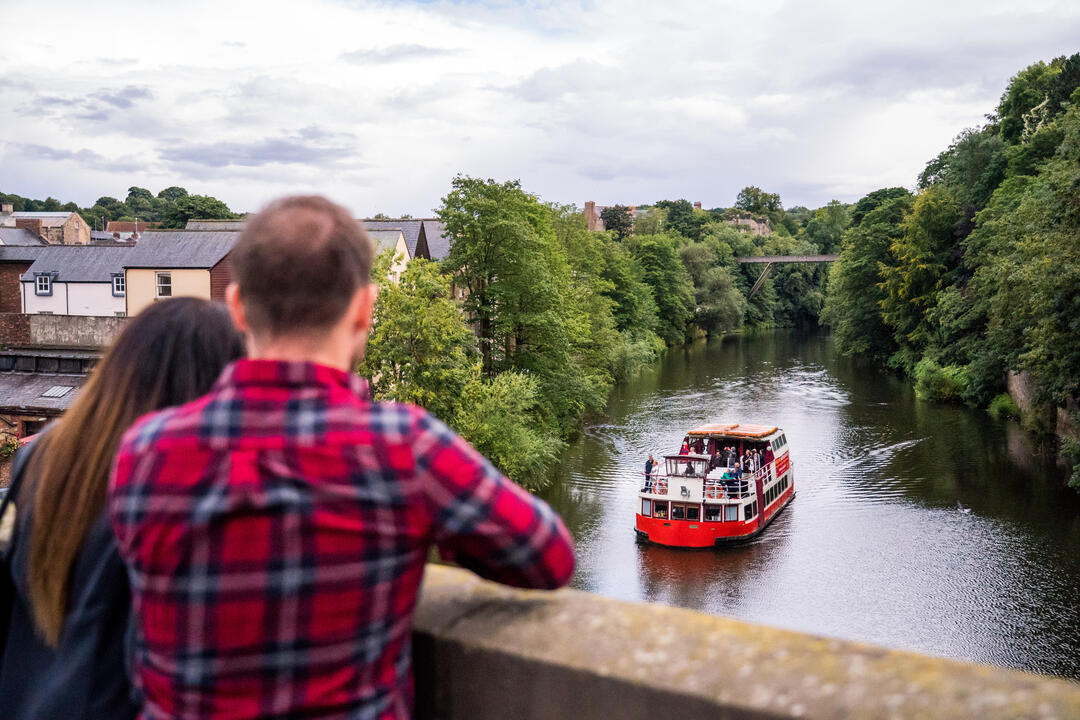 Two people stood on a bridge overlooking a tour boat sailing down a river in Durham