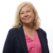 A person with blonde hair smiles at the camera, wearing a dark blazer and pink top, standing against a plain white background.
