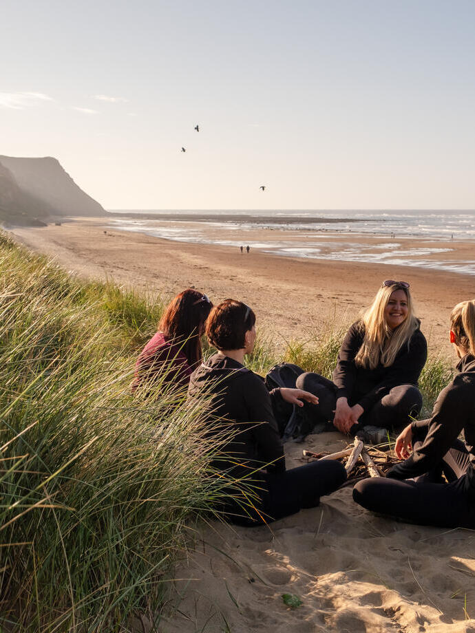 A group of female friends sit in sand dunes with a vast beach beyond