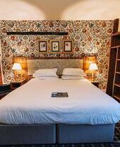 Cozy hotel room with a large bed, floral wallpaper, brick fireplace, open wardrobe, and bedside lamps creating a warm atmosphere.