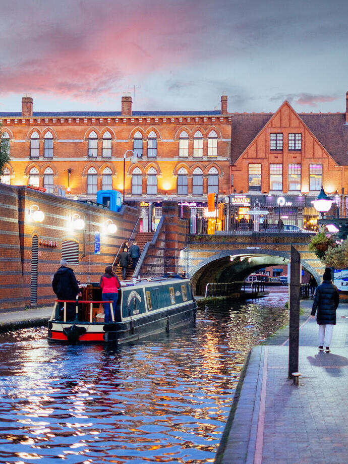 Flussboot, das in der Abenddämmerung einen Kanal hinunterfährt, während Menschen auf einem parallel verlaufenden Weg spazieren gehen.