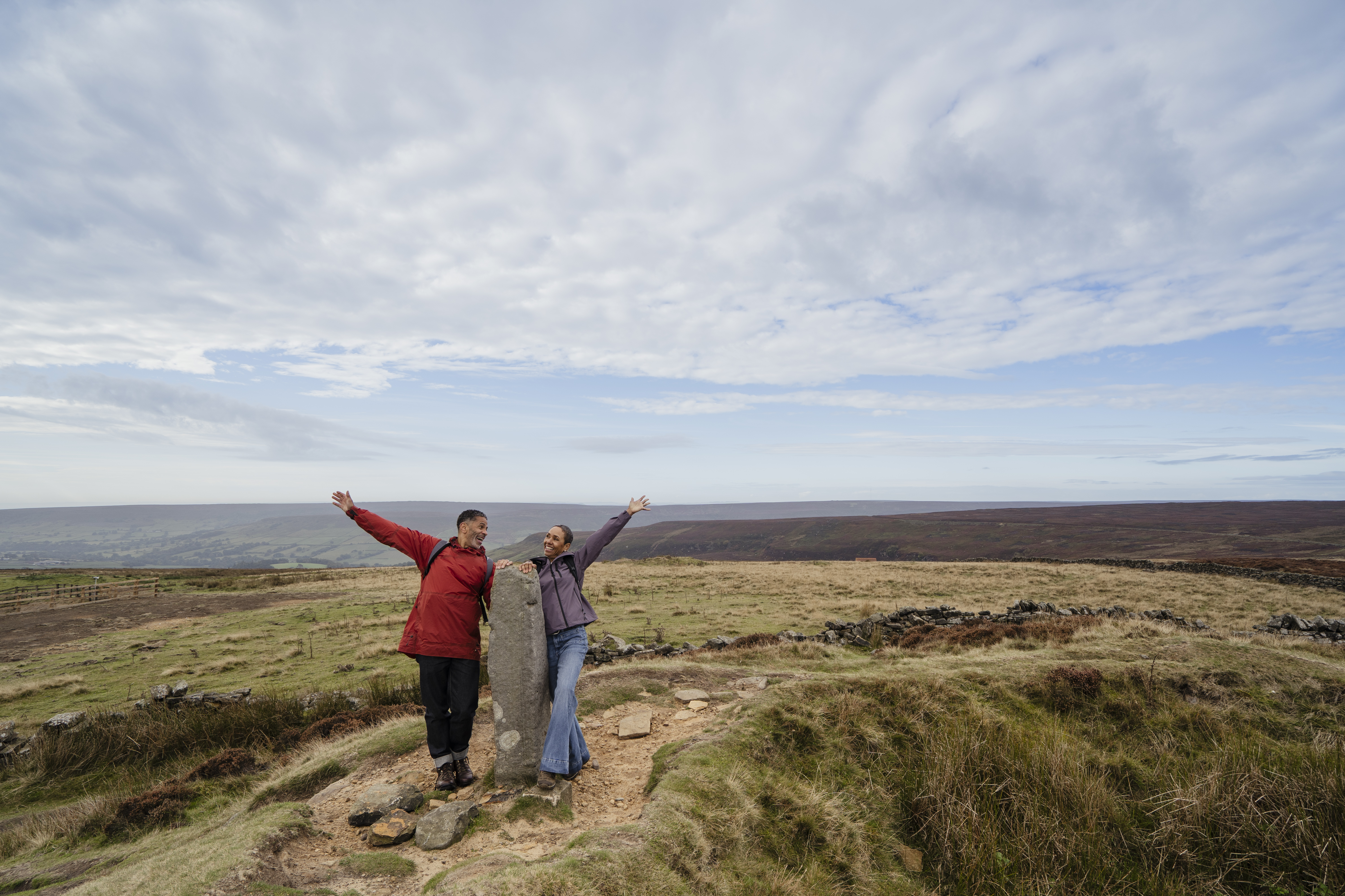 Two people posing cheerfully beside a standing stone in open countryside under a wide, partly cloudy sky. Rolling hills and grassy moorland in the background.