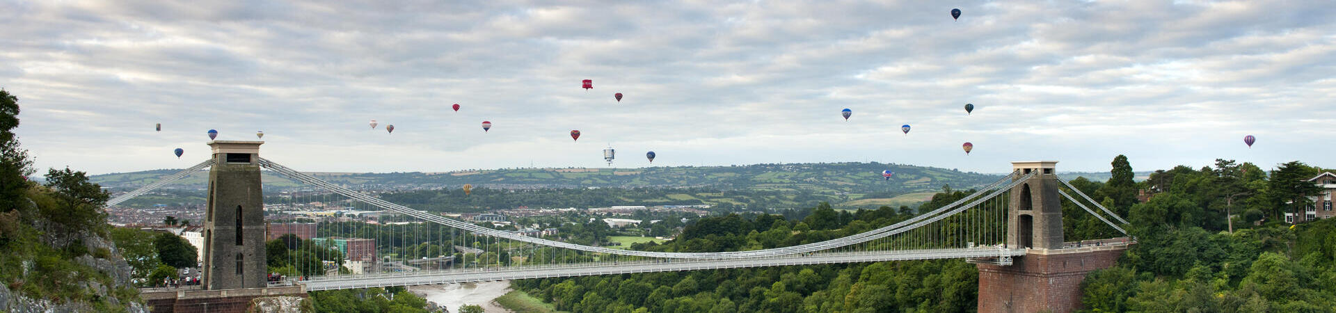 Hot air balloons floating above a river and a suspension bridge.