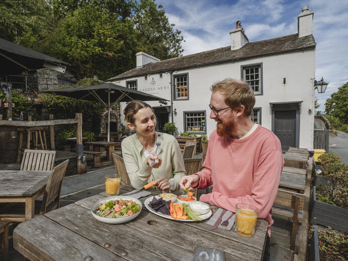 Ein Mann und eine Frau sitzen an einem Holztisch im Freien vor einem weißen Gebäude, teilen einen Teller Salat und Gemüsesnacks mit Getränken.