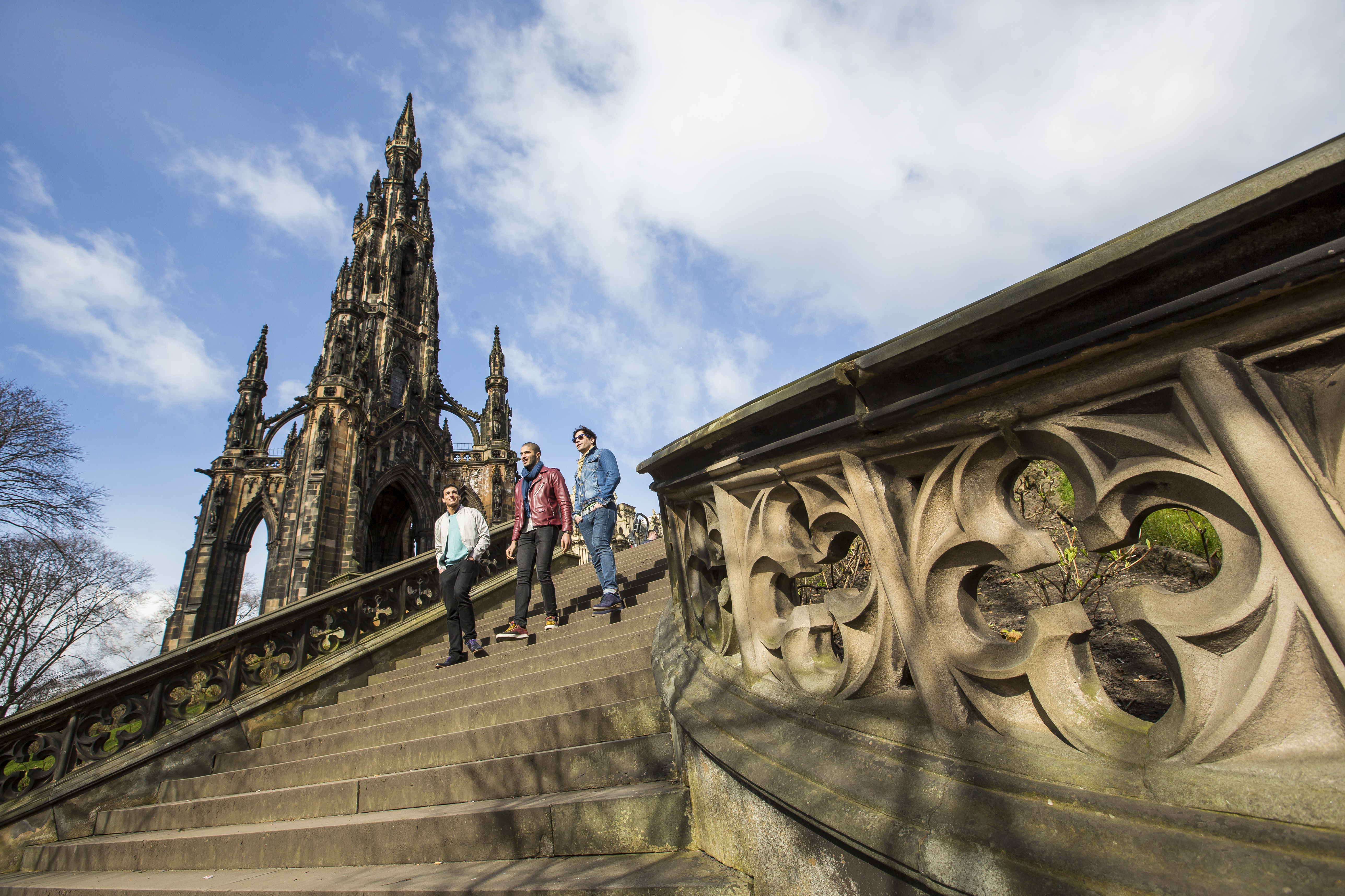Three people walking down stone steps near the ornate Scott Monument under a partly cloudy blue sky in Edinburgh.