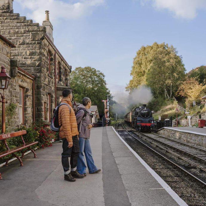 Historic railway station with stone buildings, a steam train arriving, and people waiting on the platform. Autumn trees and clear sky in the background.