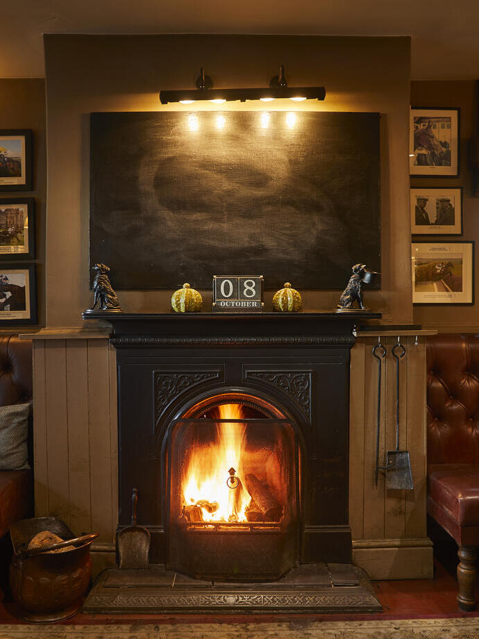 A fireplace with a lit fire flanked by two leather sofas with tables. The wall is covered in numerous framed artworks. The Duncombe Arms - Bronze award winner for the Pub of the Year at the VisitEngland Awards for Excellence 2023.