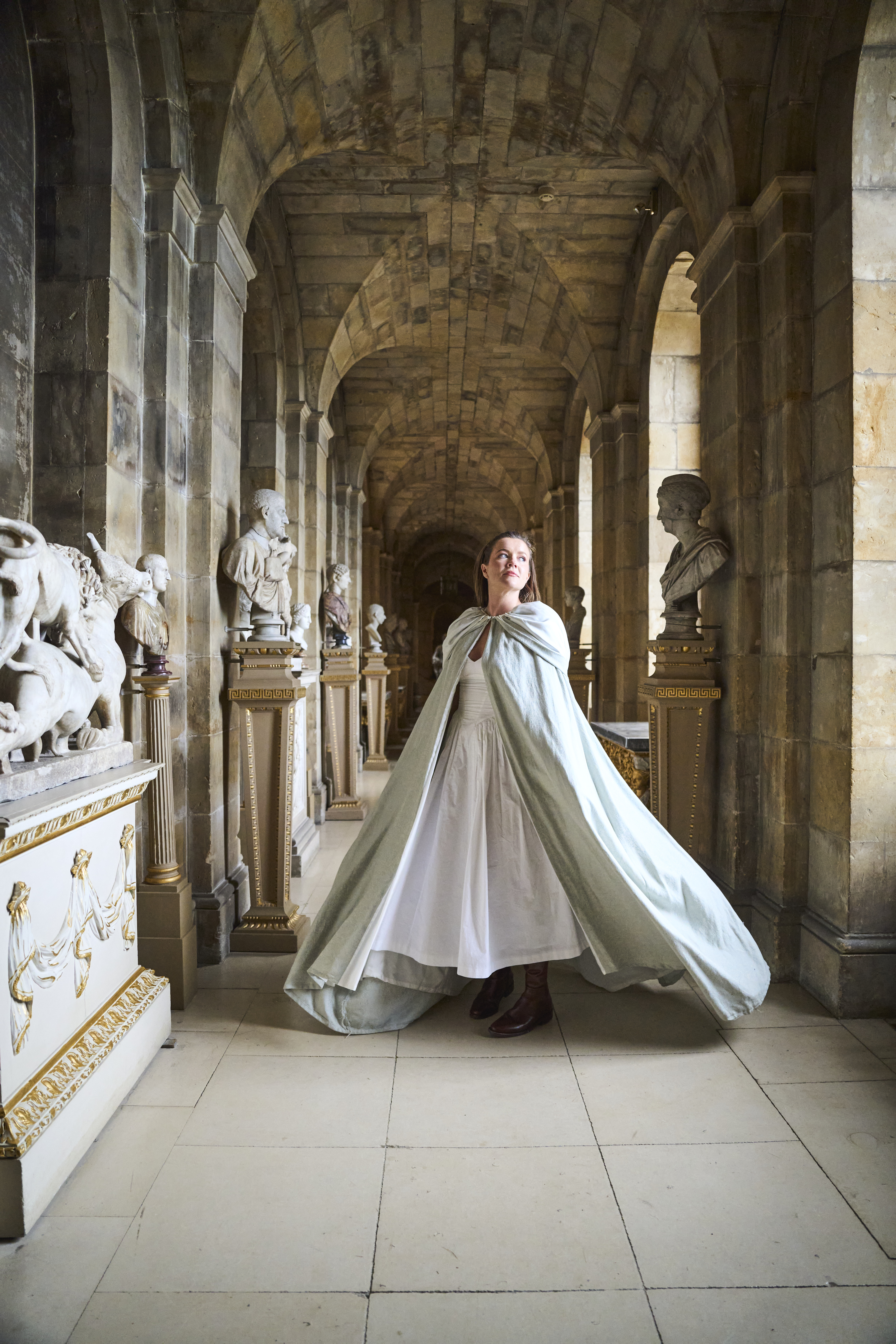 Person in flowing cloak walks through ornate stone corridor lined with classical statues and archways.