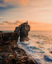 Frau steht auf einem Felsbrocken und blickt bei Sonnenuntergang aufs Meer