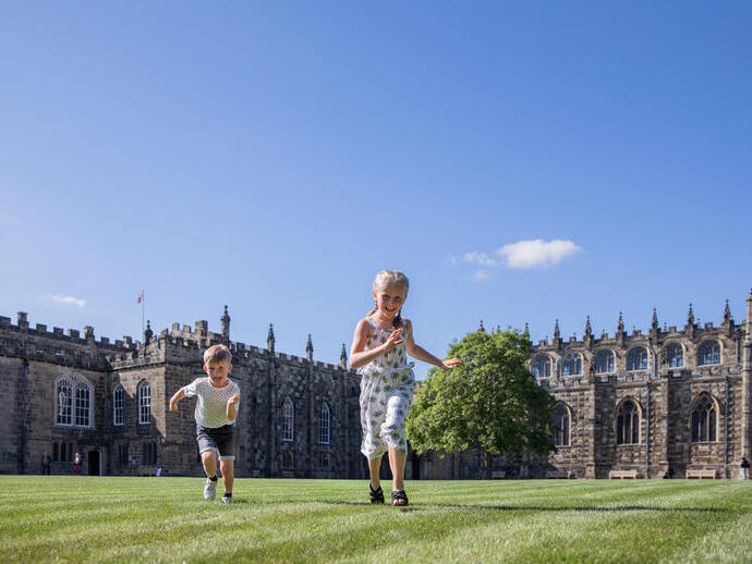 Children running and playing on grass in front of a large historic stone building under a bright blue sky.