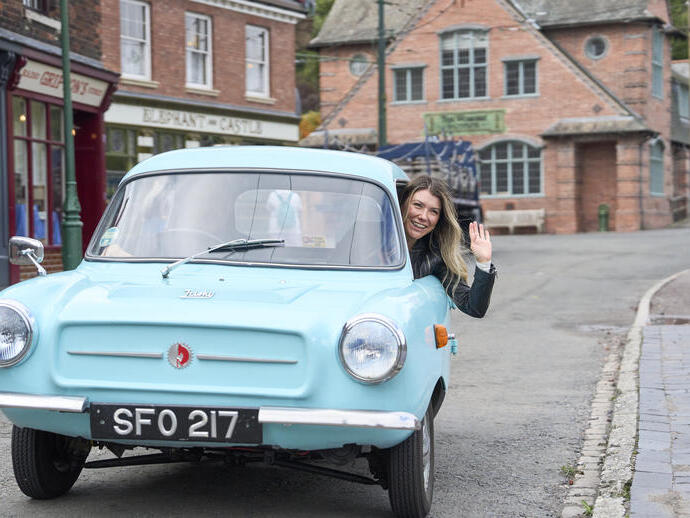A woman waves from the window of a light blue vintage car on a quiet street with historic brick buildings and shops in the background.