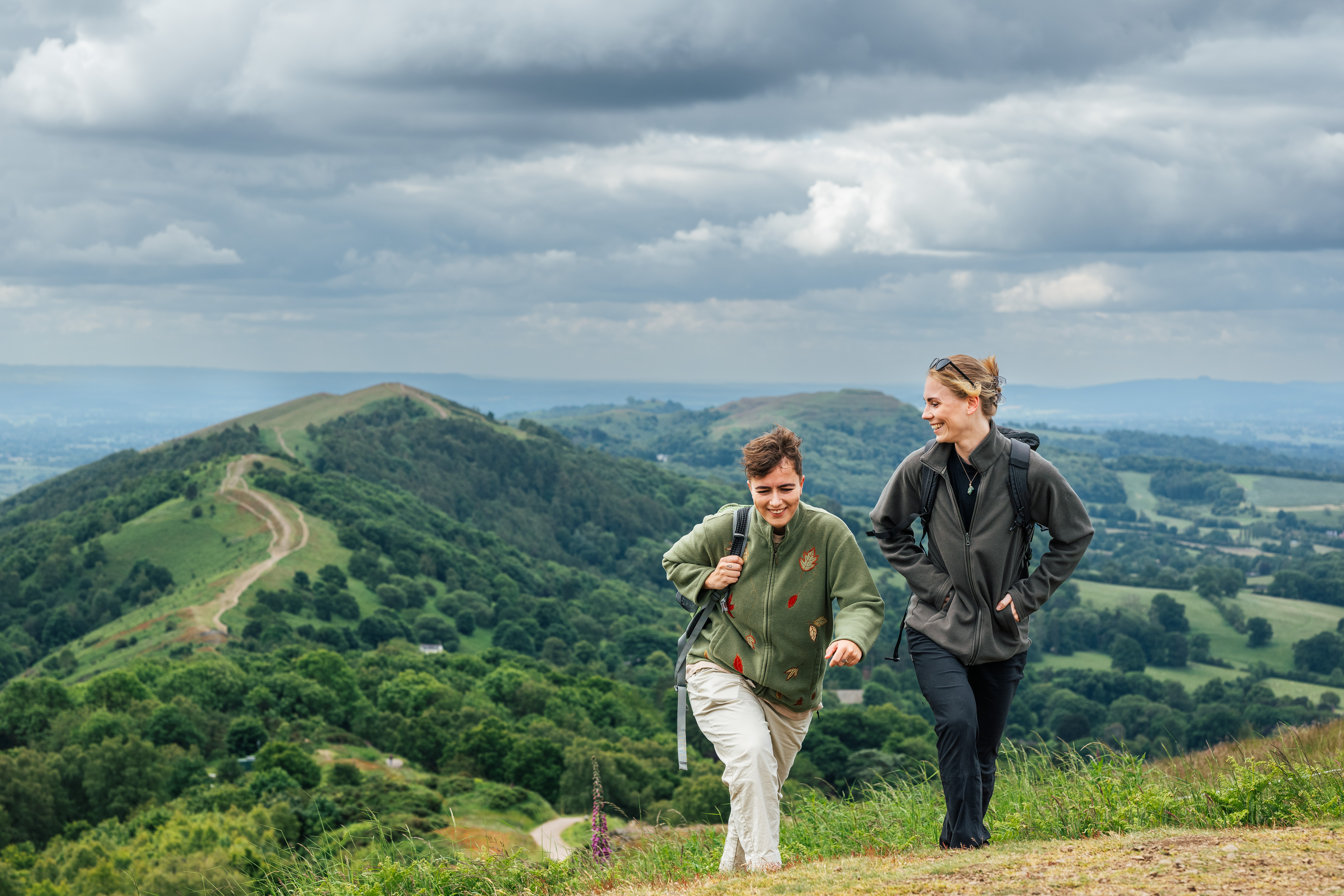Two women hike carrying rucksacks along green hills on a cloudy day
