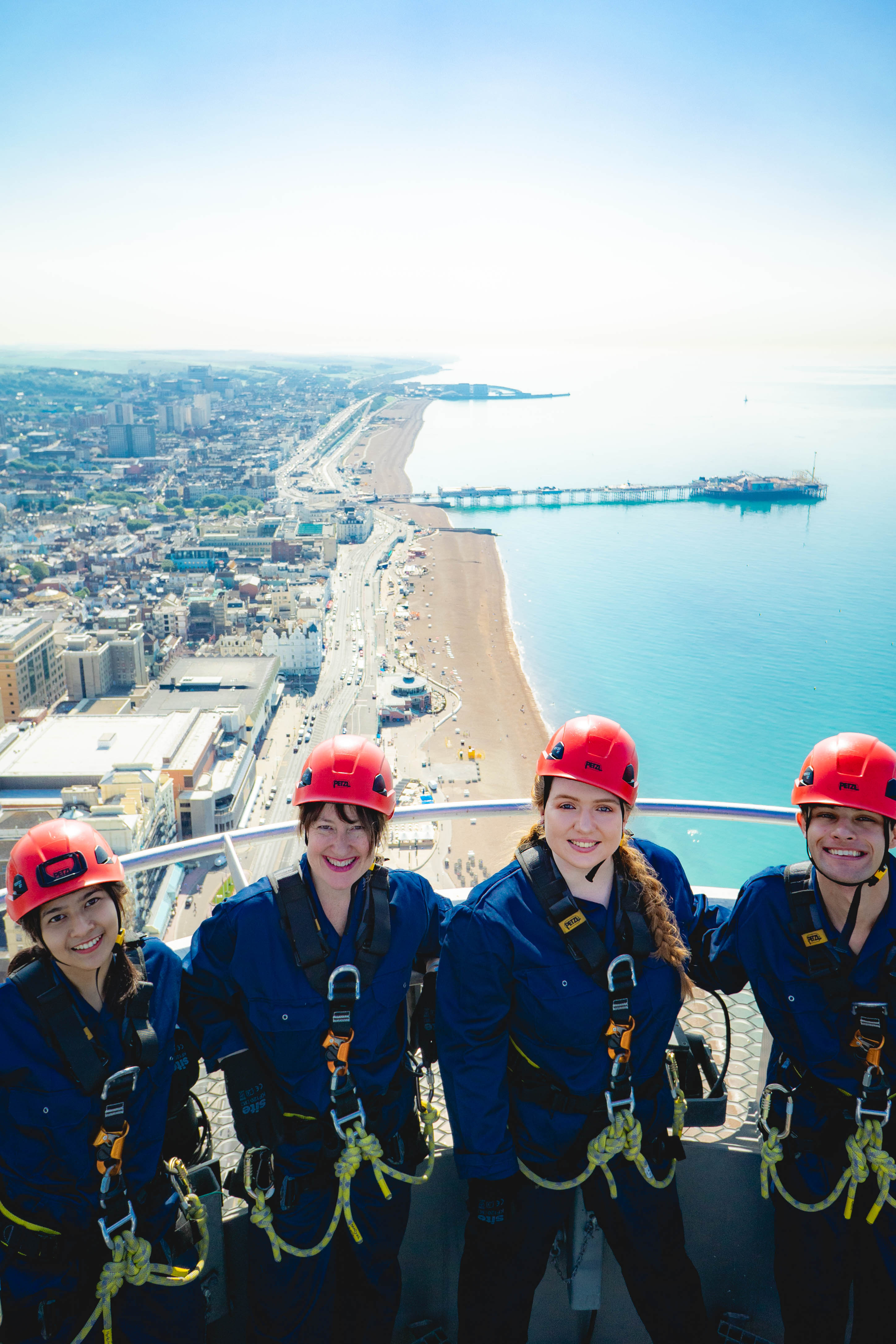 Group of friends at the top of i360 pod in climbing gear with coastline of Brighton. below