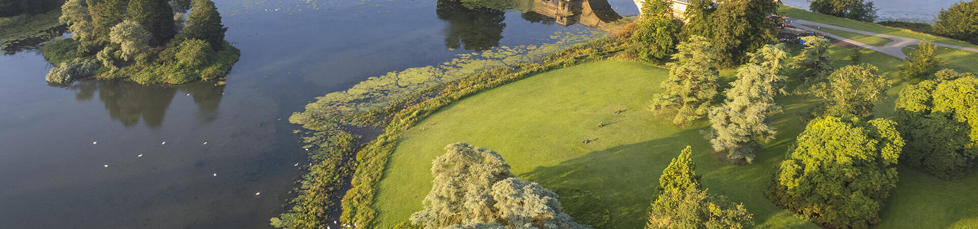 An aerial view of an eighteenth-century palace, surrounded by gardens, trees and a lake crossed by a bridge